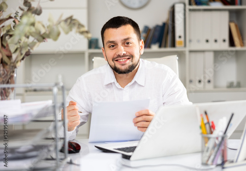 Cheerful man bookkeeper doing paperwork in his workplace in office. Smiling office manager looking at camera.
