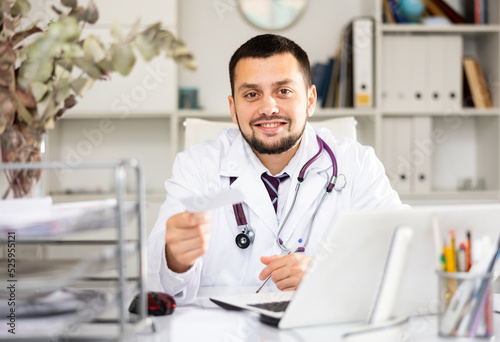 Portrait of cheerful male doctor sitting at working table and giving treatment sheet.