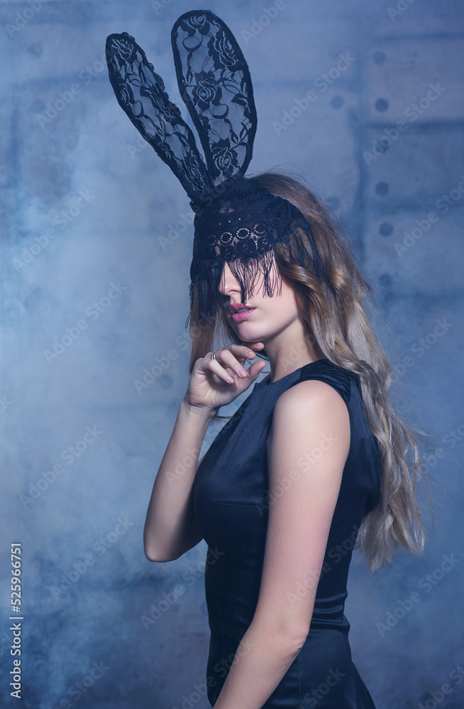 Fototapeta premium Young blonde woman in black rabbit or hare fancy mask and black dress. Female on smoke and metal wall background.