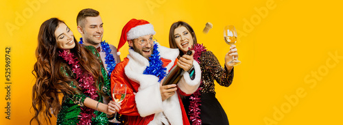 A group of young adult friends in New Year's tinsel and a santa costume shoot a champagne cork on a yellow isolated background with copy space. Banner for new year party