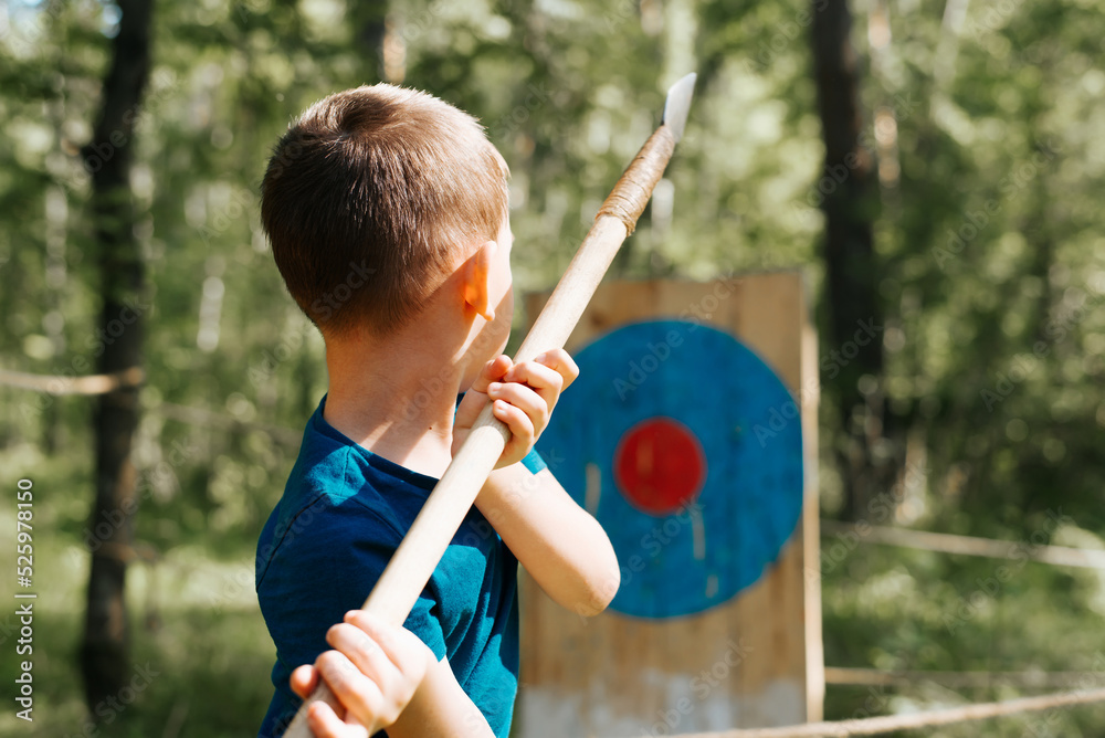 Fototapeta premium Little caucasian boy throwing a spear at a wooden target at the festival of medieval culture and outdoor games, rear view. Active childhood