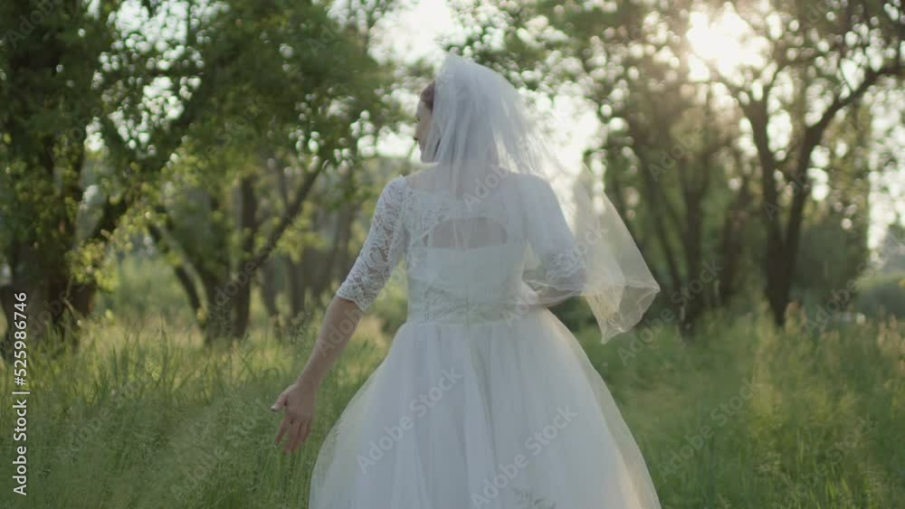 Bride in white wedding dress with bridal veil walking in the park. Newlywed touching grass. Slow motion, handheld camera, back view. Bride in white wedding dress with bridal veil walking in the park. Newlywed touching grass. Slow motion, handheld camera, back view.