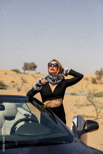 A stylish girl in a black suit and a silk scarf enjoys a ride in a convertible car in the desert, Dubai, UAE