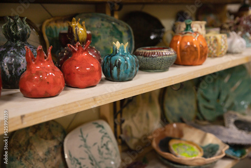 Handmade ceramic tableware on a shelf in various shapes and colors. Finished pottery hand-sculpted from clay and painted in various colors in the form of red pomegranate, plates and vases.