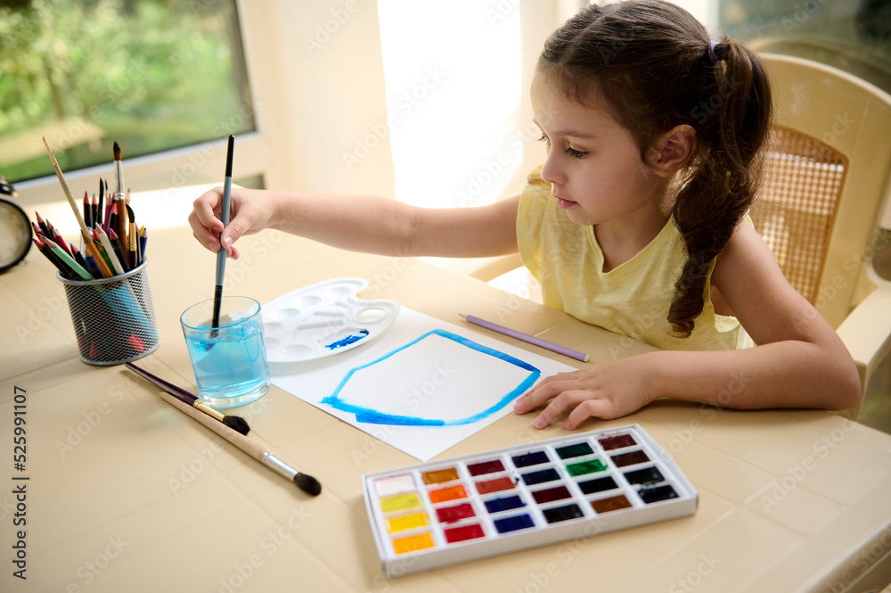 Naklejka premium Adorable Caucasian preschooler girl washes the paintbrush in a glass with water while painting picture with watercolor paints. Education, art, fun and creativity concept. Kids entertainment and hobby.