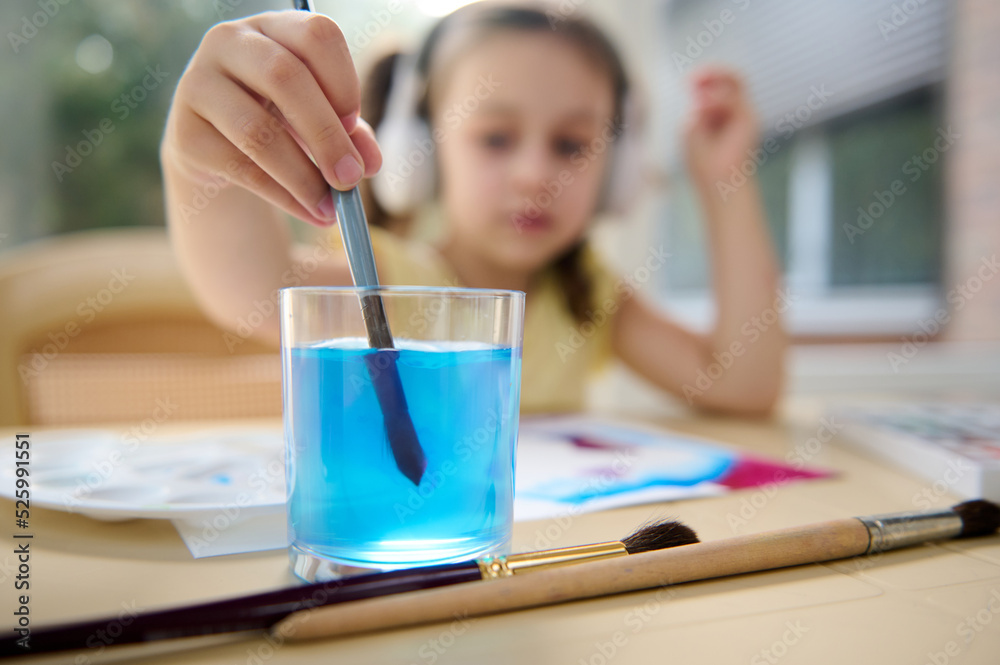 Details: hand of blurred Caucasian child, a preschool girl, holding a paintbrush and dipping it into a glass of water while painting with watercolors. Art class and courses for creative talented kids Details: hand of blurred Caucasian child, a preschool girl, holding a paintbrush and dipping it into a glass of water while painting with watercolors. Art class and courses for creative talented kids