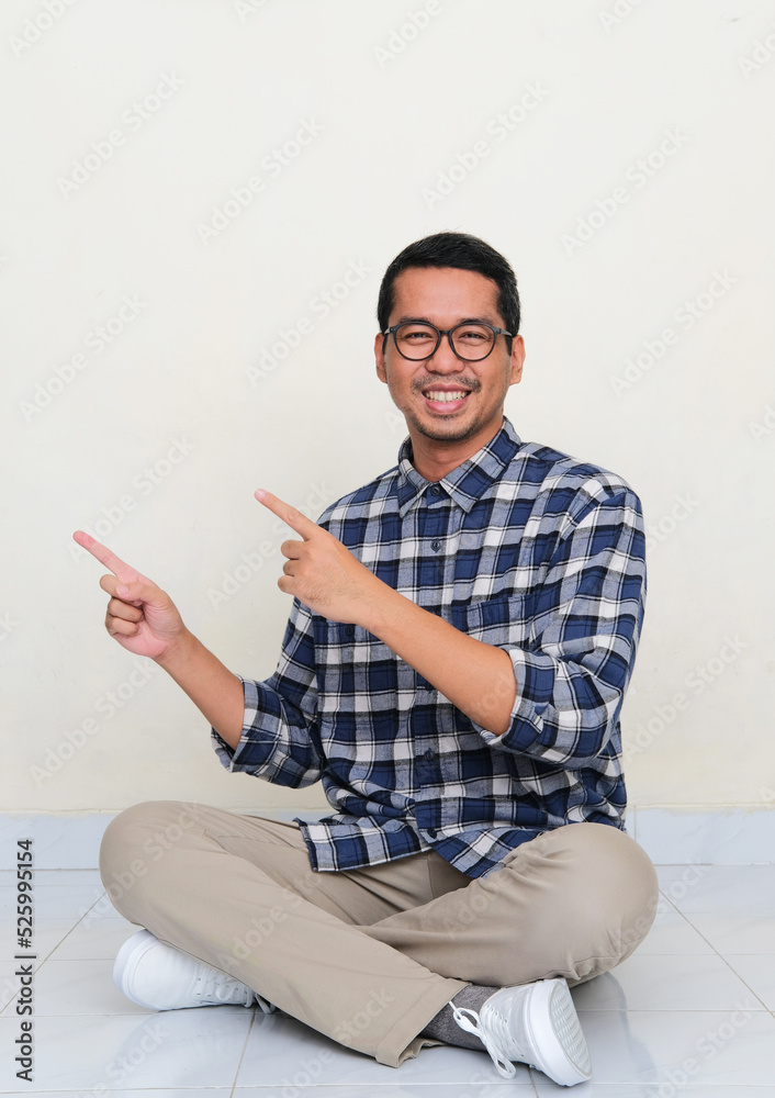 Asian man sitting in the floor smiling to camera while pointing to the right side Asian man sitting in the floor smiling to camera while pointing to the right side