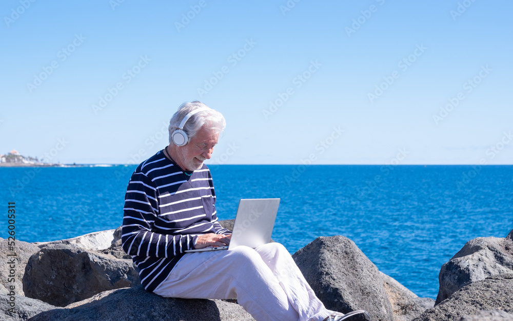 Smiling senior caucasian man with headphones sitting outdoors at sea using laptop in remote working, white-haired elderly male browsing with computer