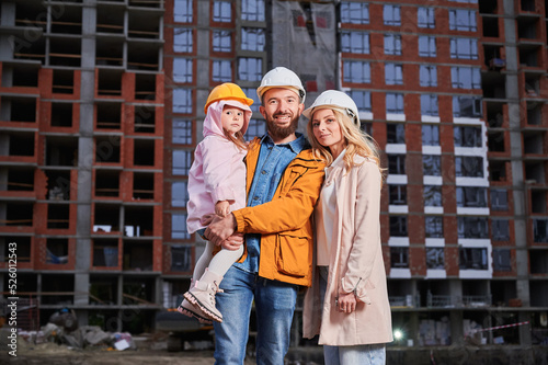 Parents and daughter smiling while standing outdoors against apartment building under construction. Portrait of happy family homeowners at construction site.
