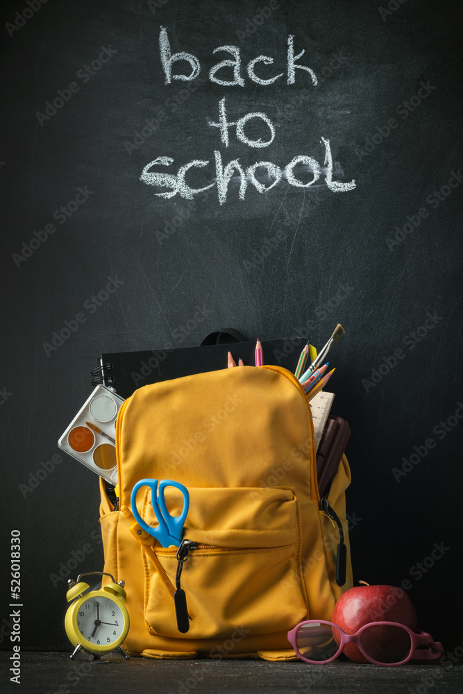 Naklejka premium Yellow backpack with school supplies next to the globe, red apple and glasses on the black school board background. Back to school concept on September day