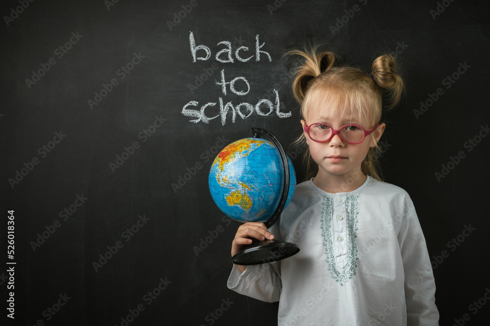 Fototapeta premium Portrait of little caucasian girl wearing glasses. Elementary school student holding the globe on the black school board background. Back to school concept