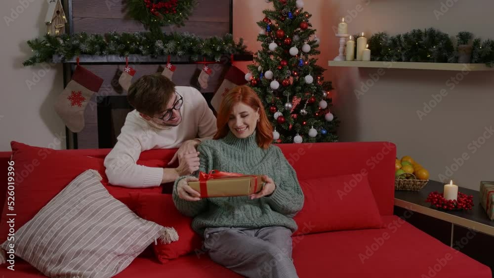 Boyfriend giving present to his loved girlfriend, celebrating New Year. Couple sitting on sofa and congratulating with gifts. Man and woman hugging, happy young family, making surprise. Boyfriend giving present to his loved girlfriend, celebrating New Year. Couple sitting on sofa and congratulating with gifts. Man and woman hugging, happy young family, making surprise.