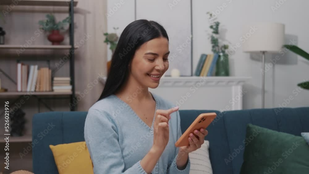 Portrait of a beautiful young woman sitting on a blue sofa and using a smartphone smiling turning her head looking at the camera in a modern room. Tapping, scrolling. Slow motion Portrait of a beautiful young woman sitting on a blue sofa and using a smartphone smiling turning her head looking at the camera in a modern room. Tapping, scrolling. Slow motion