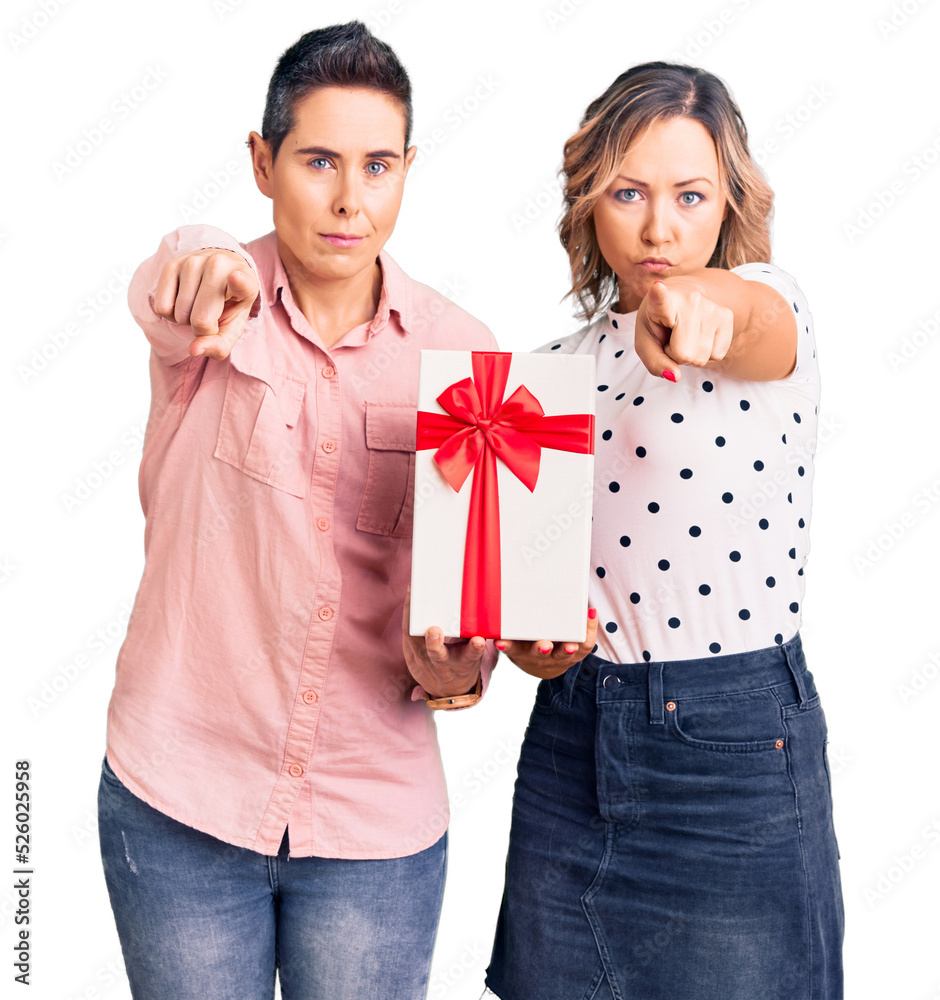 Couple of women holding gift pointing with finger to the camera and to you, confident gesture looking serious Couple of women holding gift pointing with finger to the camera and to you, confident gesture looking serious