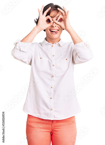 Beautiful young woman with short hair wearing elegant white shirt doing ok gesture like binoculars sticking tongue out, eyes looking through fingers. crazy expression.