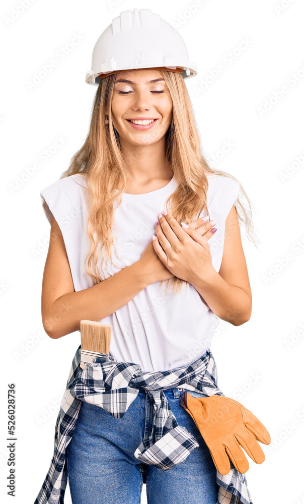 Beautiful caucasian woman with blonde hair wearing hardhat and painter clothes smiling with hands on chest with closed eyes and grateful gesture on face. health concept.