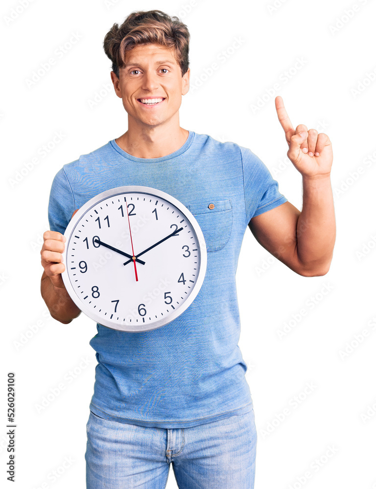 Young handsome man holding big clock surprised with an idea or question pointing finger with happy face, number one Young handsome man holding big clock surprised with an idea or question pointing finger with happy face, number one