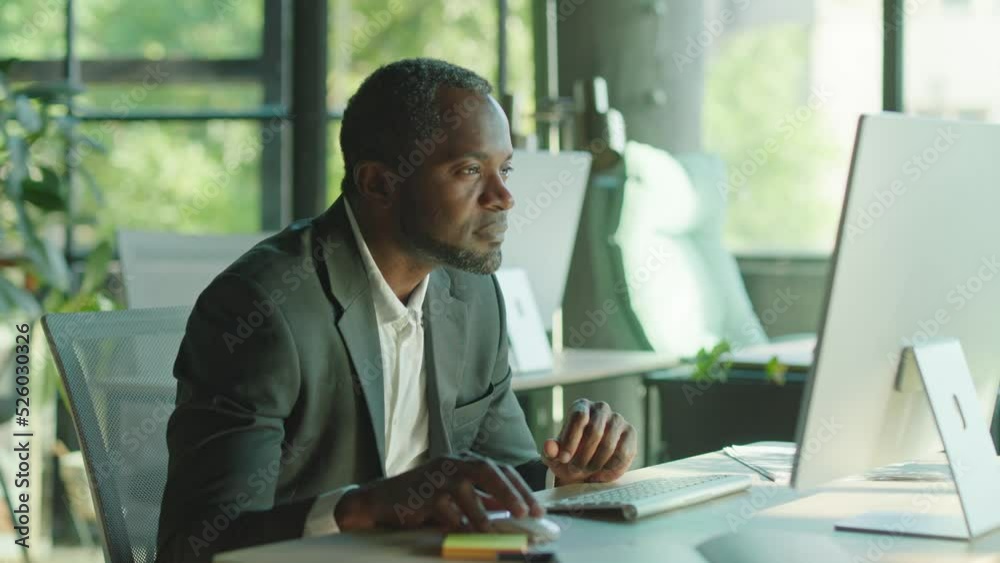 Happy attractive african american man working on the computer smiling in the office at his desk. Sunlight. Businessman. Corporate. CEO. Slow motion Happy attractive african american man working on the computer smiling in the office at his desk. Sunlight. Businessman. Corporate. CEO. Slow motion