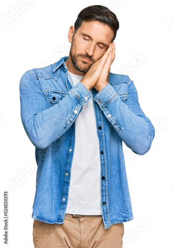 Young handsome man wearing casual denim jacket sleeping tired dreaming and posing with hands together while smiling with closed eyes.