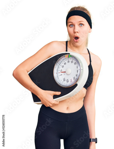 Young beautiful blonde woman wearing sportswear holding weighing machine scared and amazed with open mouth for surprise  disbelief face