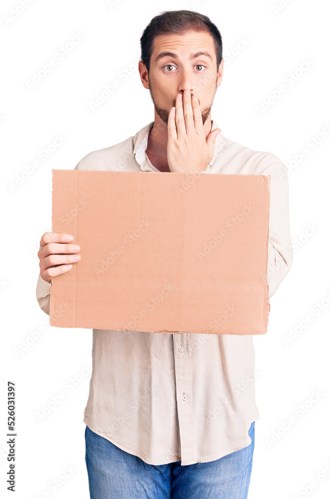 Young handsome caucasian man holding empty cardboard banner covering mouth with hand, shocked and afraid for mistake. surprised expression Young handsome caucasian man holding empty cardboard banner covering mouth with hand, shocked and afraid for mistake. surprised expression