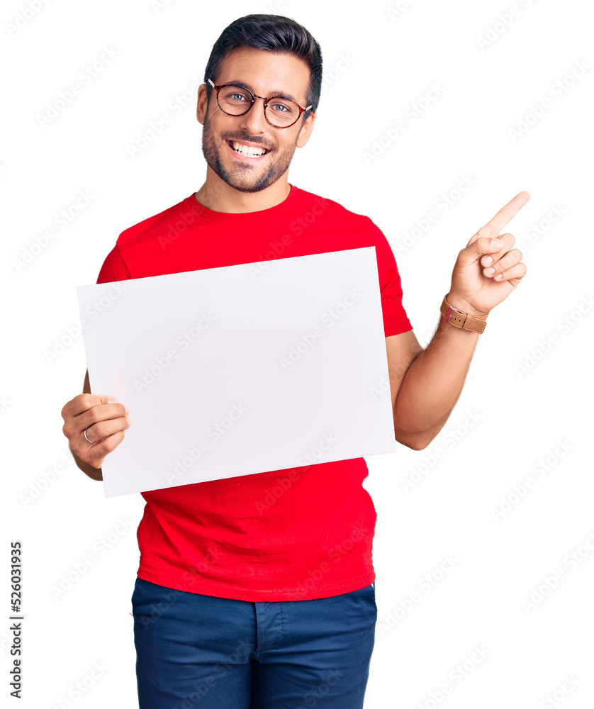 Young hispanic man holding blank empty banner smiling happy pointing with hand and finger to the side Young hispanic man holding blank empty banner smiling happy pointing with hand and finger to the side