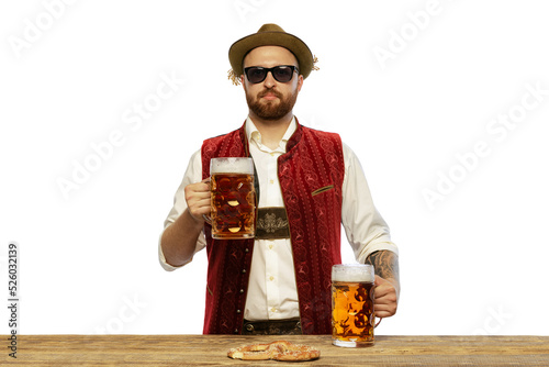 Portrait of young man wearing traditional Bavarian or German clothes and sunglasses, holding foamy beer mugs isolated over white background
