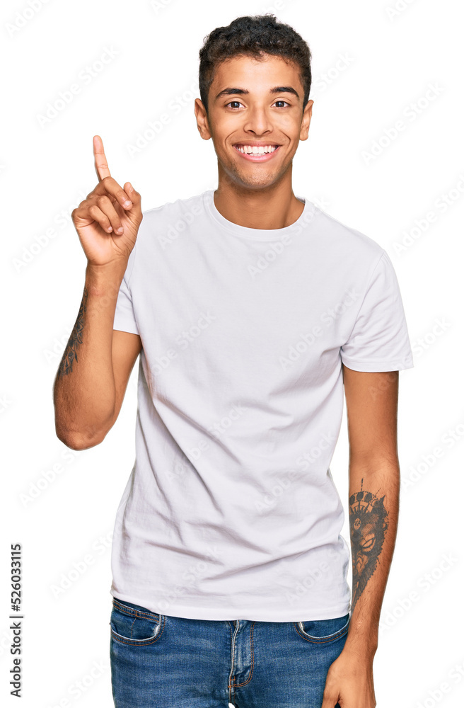Young handsome african american man wearing casual white tshirt showing and pointing up with finger number one while smiling confident and happy. Young handsome african american man wearing casual white tshirt showing and pointing up with finger number one while smiling confident and happy.