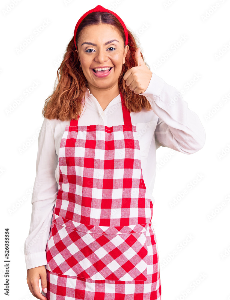 Young latin woman wearing apron smiling happy and positive, thumb up doing excellent and approval sign Young latin woman wearing apron smiling happy and positive, thumb up doing excellent and approval sign