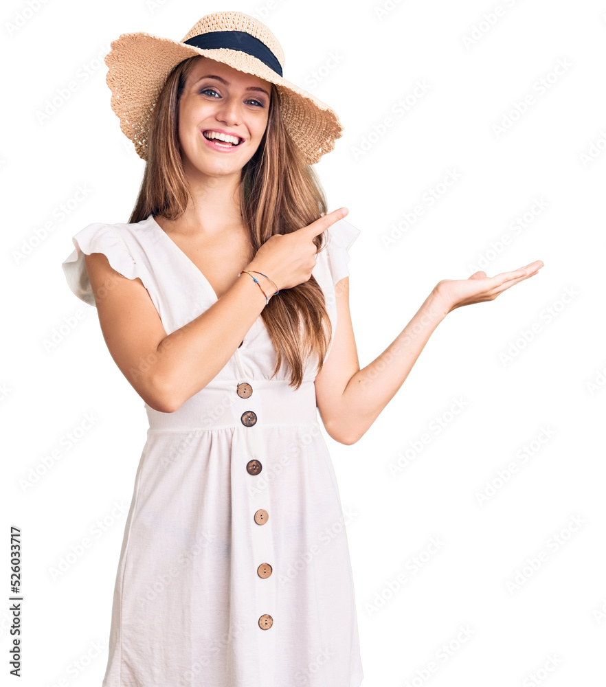 Young beautiful blonde woman wearing summer dress and hat amazed and smiling to the camera while presenting with hand and pointing with finger. Young beautiful blonde woman wearing summer dress and hat amazed and smiling to the camera while presenting with hand and pointing with finger.