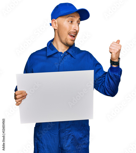 Bald man with beard wearing builder jumpsuit uniform holding empty banner pointing thumb up to the side smiling happy with open mouth