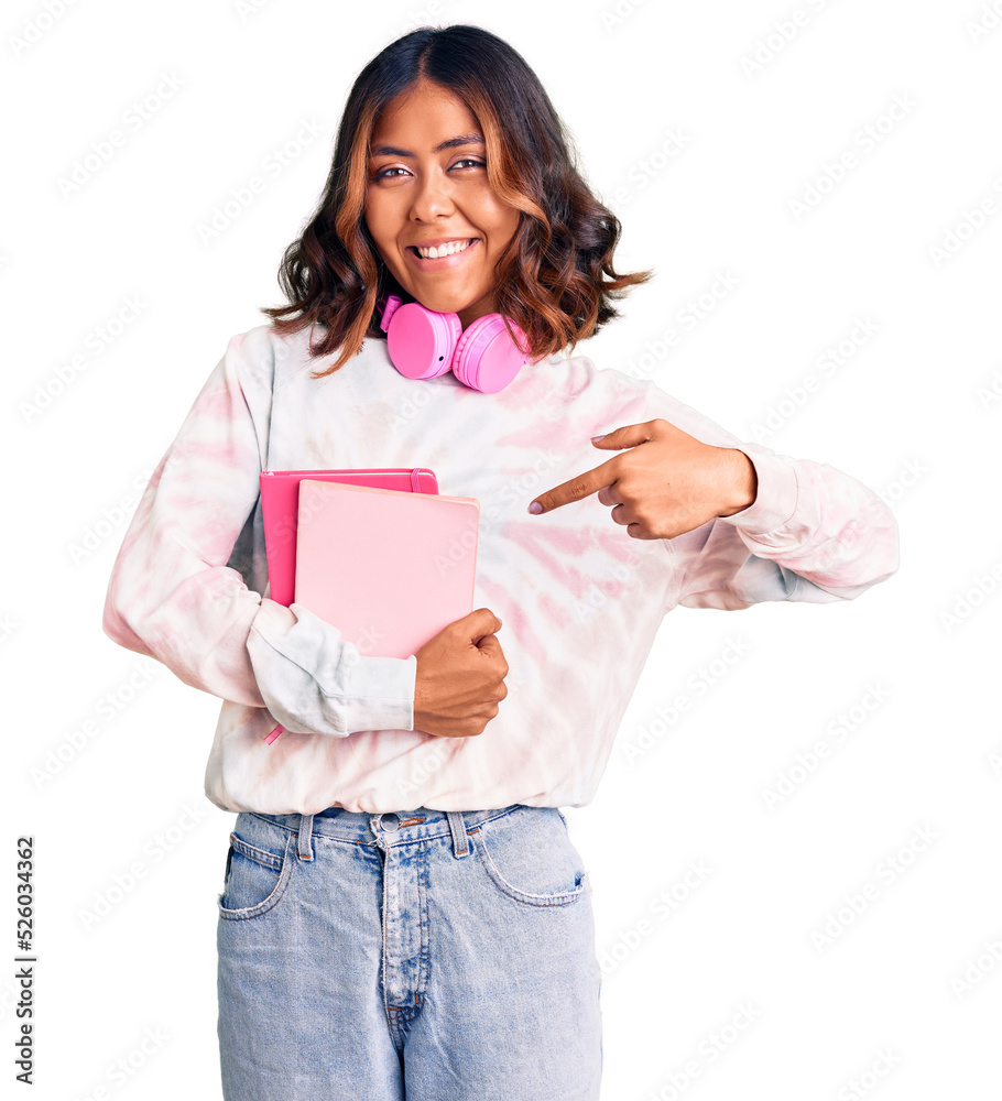 Young beautiful mixed race woman holding books wearing headphones smiling happy pointing with hand and finger Young beautiful mixed race woman holding books wearing headphones smiling happy pointing with hand and finger