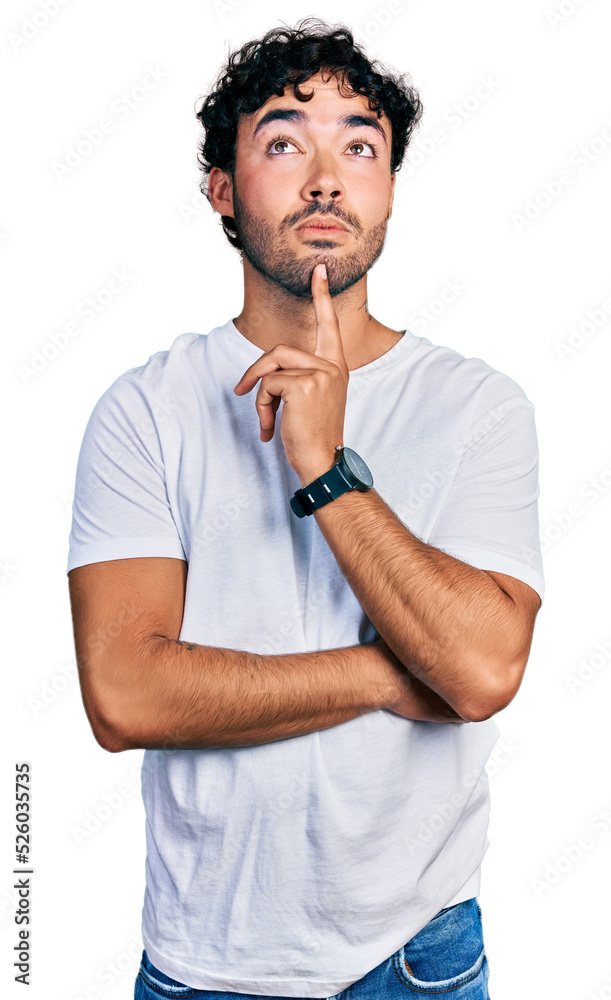 Hispanic young man with beard wearing casual white t shirt thinking concentrated about doubt with finger on chin and looking up wondering Hispanic young man with beard wearing casual white t shirt thinking concentrated about doubt with finger on chin and looking up wondering