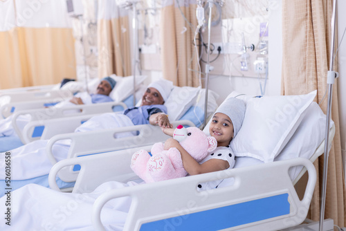 Portrait Of Girl Lying In Bed In Hospital Ward Hugging Teddy Bear