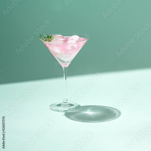Tropical cold drink cocktail minimal concept with pastel pink martini glass and ice on green color background. Trendy sunlight summer party aesthetic.
