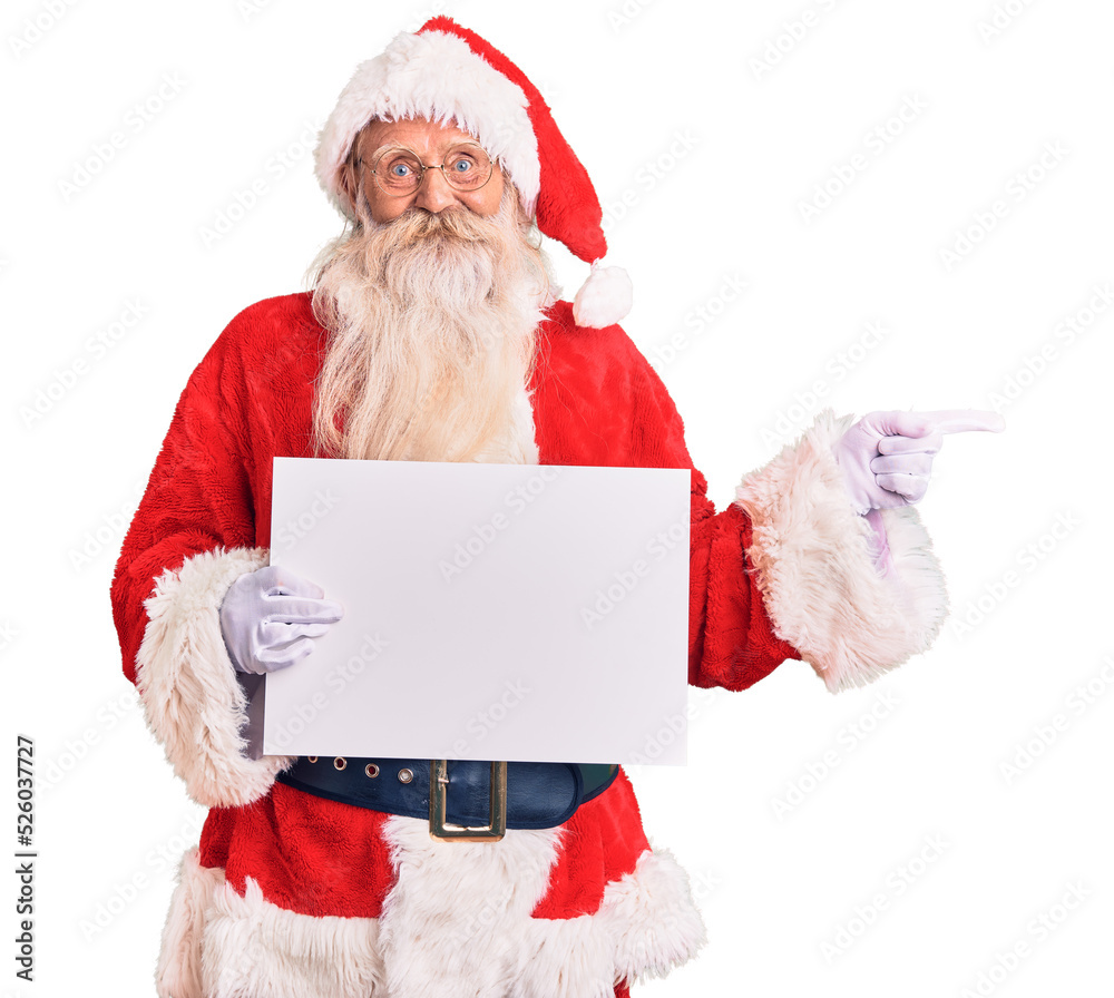 Old senior man with grey hair and long beard wearing santa claus costume holding banner smiling happy pointing with hand and finger to the side Old senior man with grey hair and long beard wearing santa claus costume holding banner smiling happy pointing with hand and finger to the side