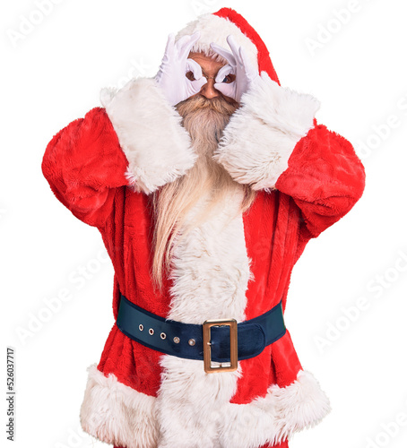 Old senior man with grey hair and long beard wearing traditional santa claus costume doing ok gesture like binoculars sticking tongue out, eyes looking through fingers. crazy expression.