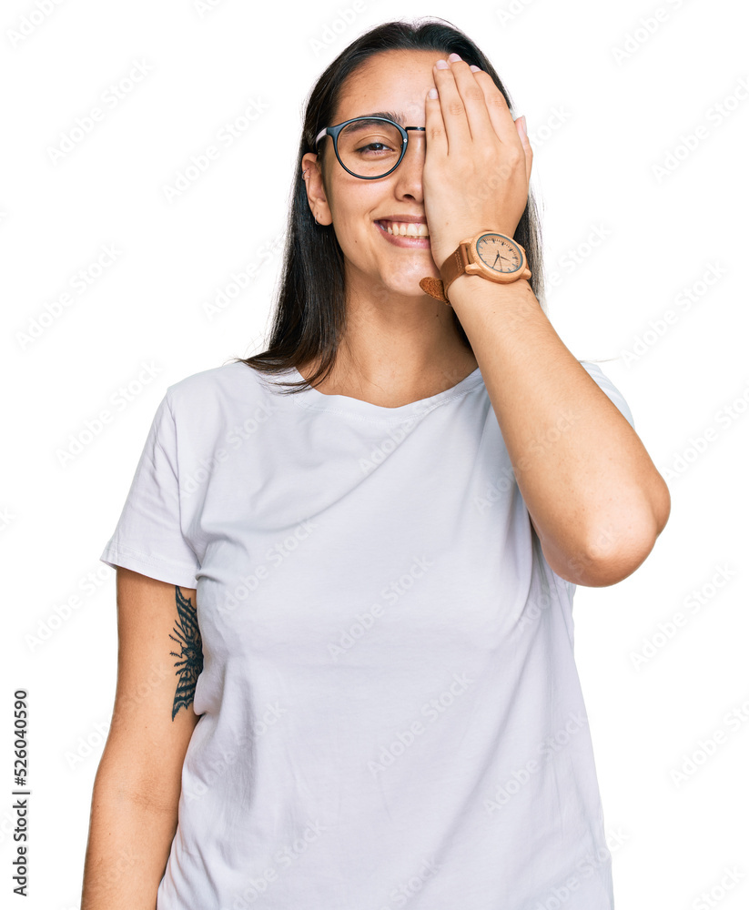 Young hispanic woman wearing casual white t shirt covering one eye with hand, confident smile on face and surprise emotion. Young hispanic woman wearing casual white t shirt covering one eye with hand, confident smile on face and surprise emotion.