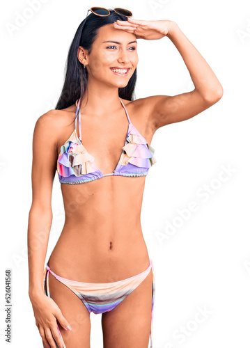 Young beautiful latin girl wearing bikini and sunglasses very happy and smiling looking far away with hand over head. searching concept.