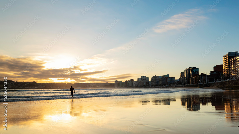 Beautiful panoramic view of the sunrise over the bay of San Lorenzo in Gijón, Spain. Sunrise over Gijón with reflections of the sun on the wet sand. Backlit silhouette in Gijón