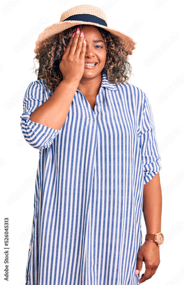 Young african american plus size woman wearing summer hat covering one eye with hand, confident smile on face and surprise emotion.