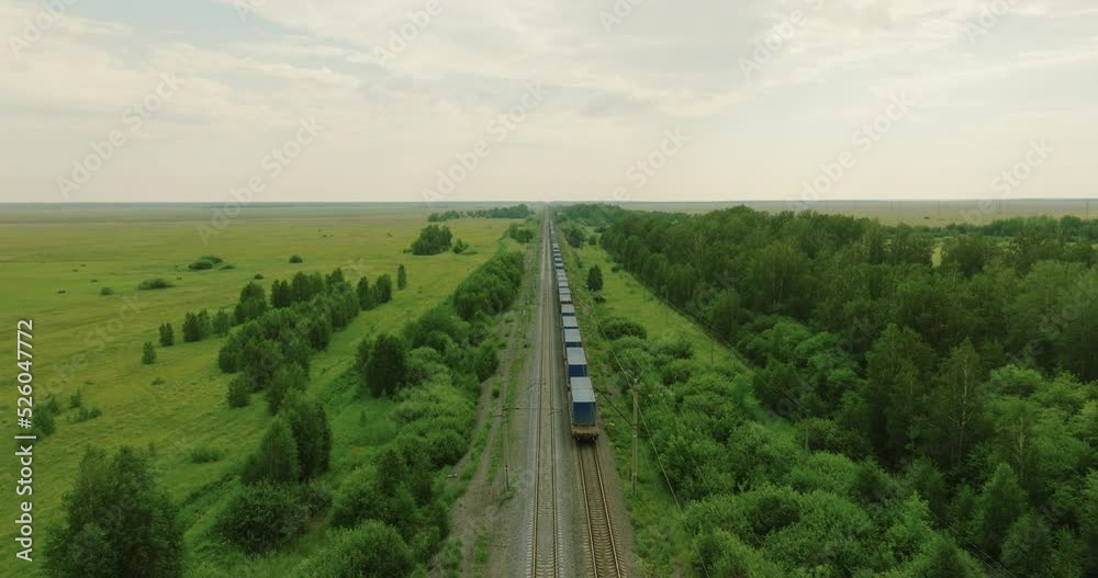 Freight long train carries with cargo carriages in flat swampland landscape through a difficult part of  Siberian railways. Aerial drone view at summer sunset