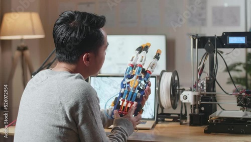 Wallpaper Mural Side View Of Asian Male With 3D Printing Checking A Cyborg Hand While Designing It On A Laptop At Home
 Torontodigital.ca