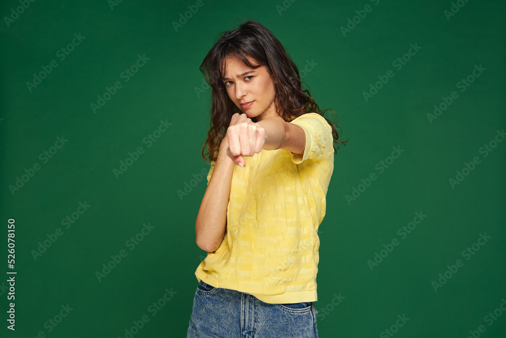 Serious confident young girl punching with clenched fist at camera, frowning on green background. Boxing, battle concept