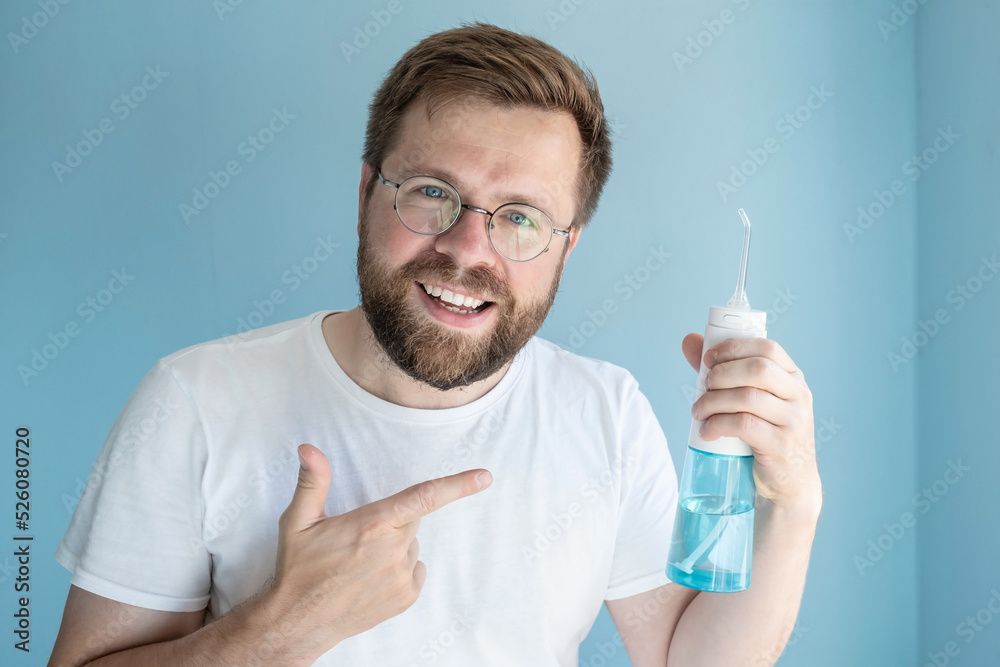 Cute smiling man holds a portable irrigator in hand, he recommends this oral hygiene product and points to it with his finger Cute smiling man holds a portable irrigator in hand, he recommends this oral hygiene product and points to it with his finger