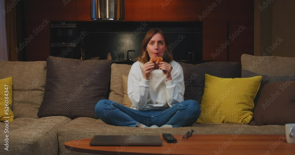 Young woman watching television series or film eating burger, sitting on sofa at home at night on weekend Young woman watching television series or film eating burger, sitting on sofa at home at night on weekend