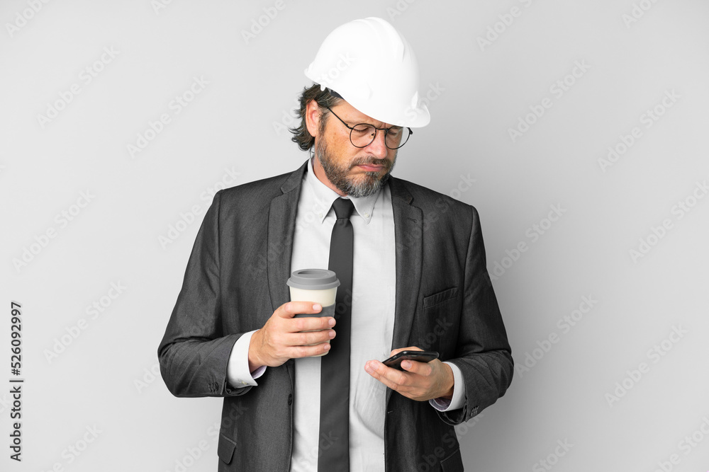Young architect man with helmet over isolated background holding coffee to take away and a mobile