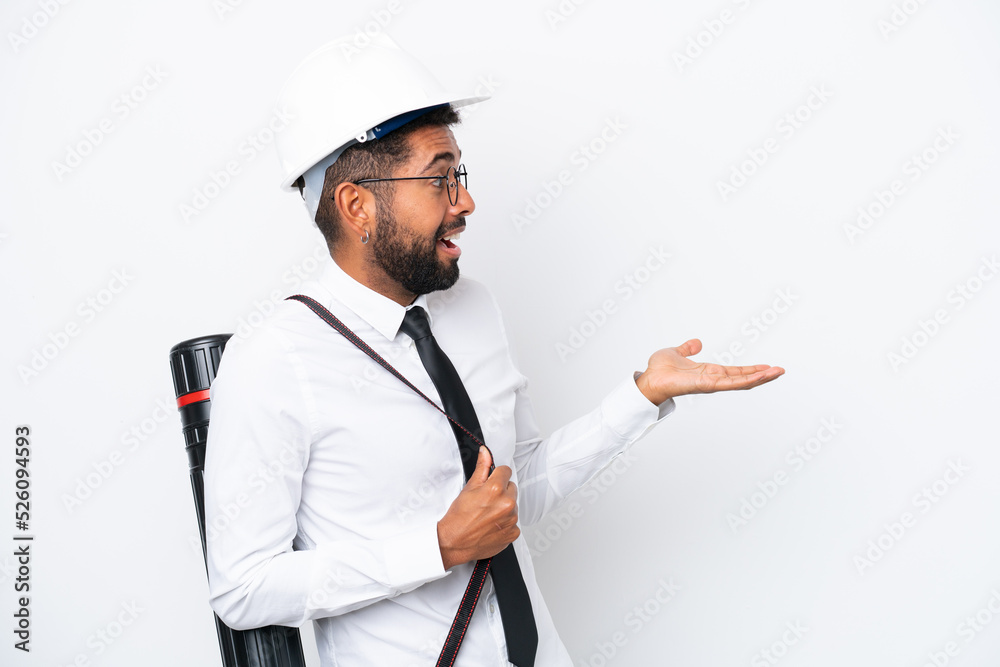 Young architect Brazilian man with helmet and holding blueprints isolated on white background with surprise expression while looking side Young architect Brazilian man with helmet and holding blueprints isolated on white background with surprise expression while looking side