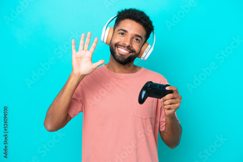 Young brazilian man playing with a video game controller isolated on blue background counting five with fingers