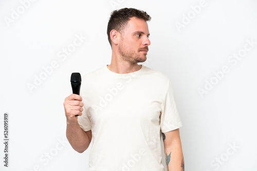 Young caucasian singer man picking up a microphone isolated on white background looking to the side